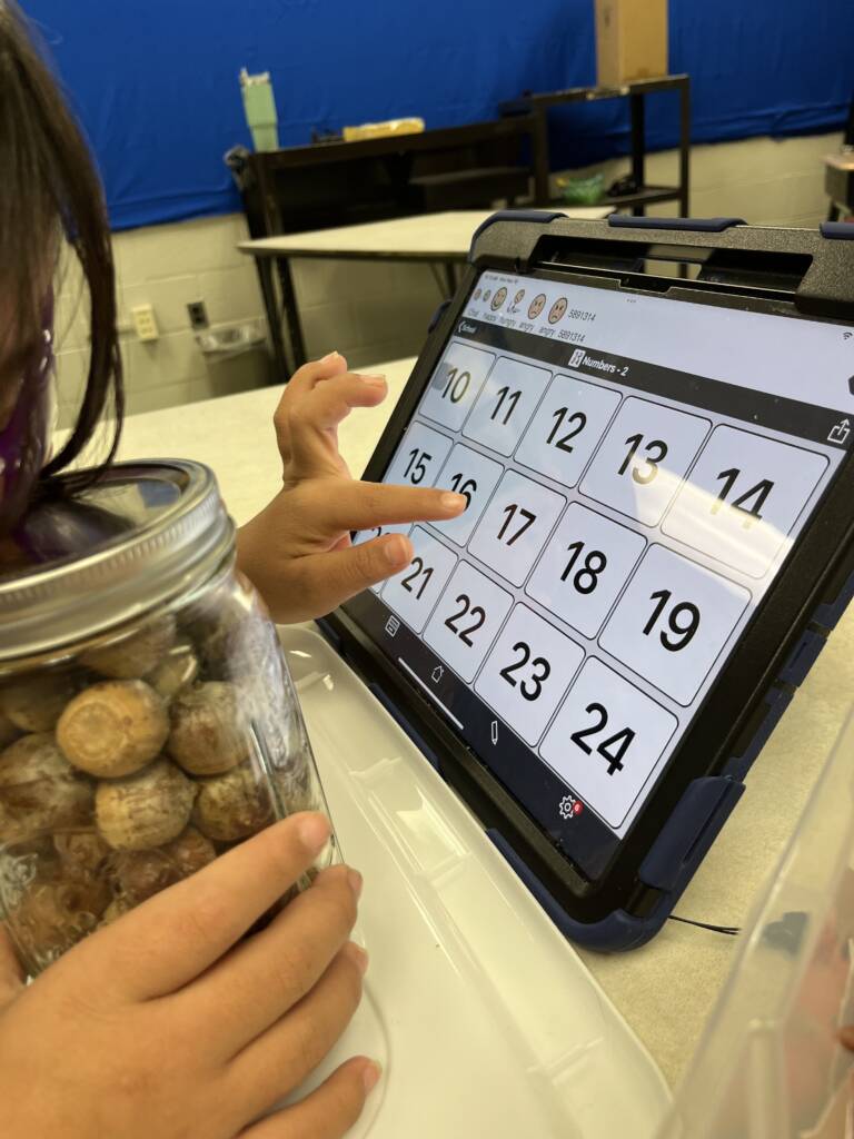 Student using her talker to find numbers so she can guess how many candy corns are in a jar. 