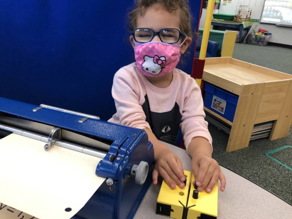 A young girl with a mask on poses with a brailler.