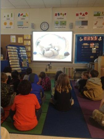 Children sitting on classroom floor looking at screen in the front of the classroom
