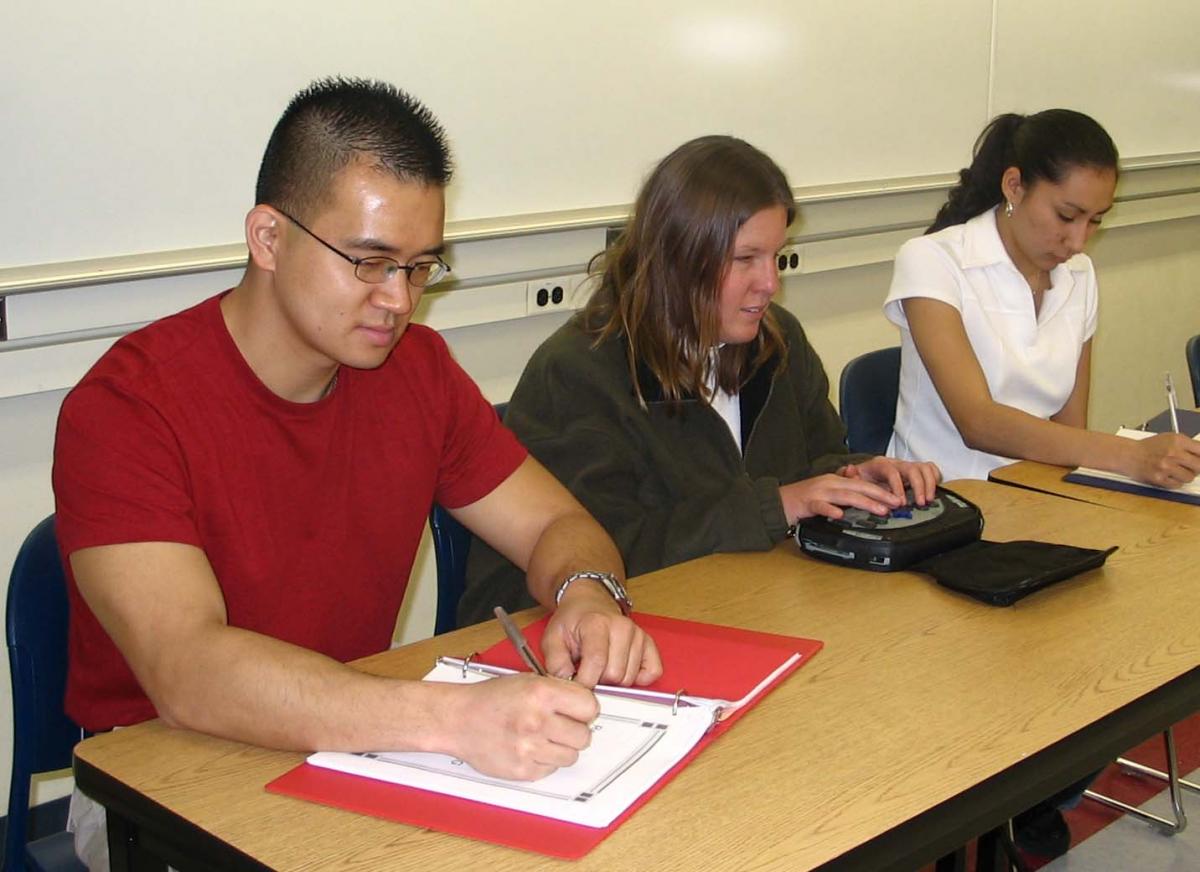 Anna sits between a male student and a female student.  All 3 are taking notes - Anna with her portable note taker and the others with pen and paper.