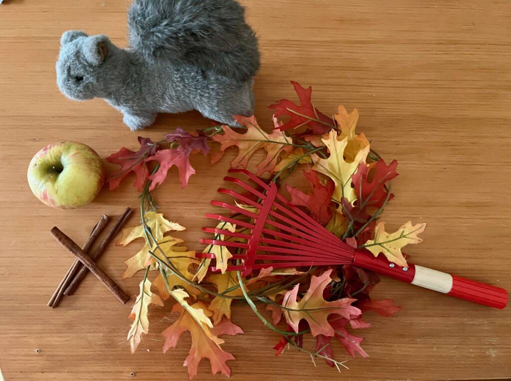 Autumn items that include an apple, plastic leaves, cinnamon sticks, rake, and a squirrel stuffed animal. 