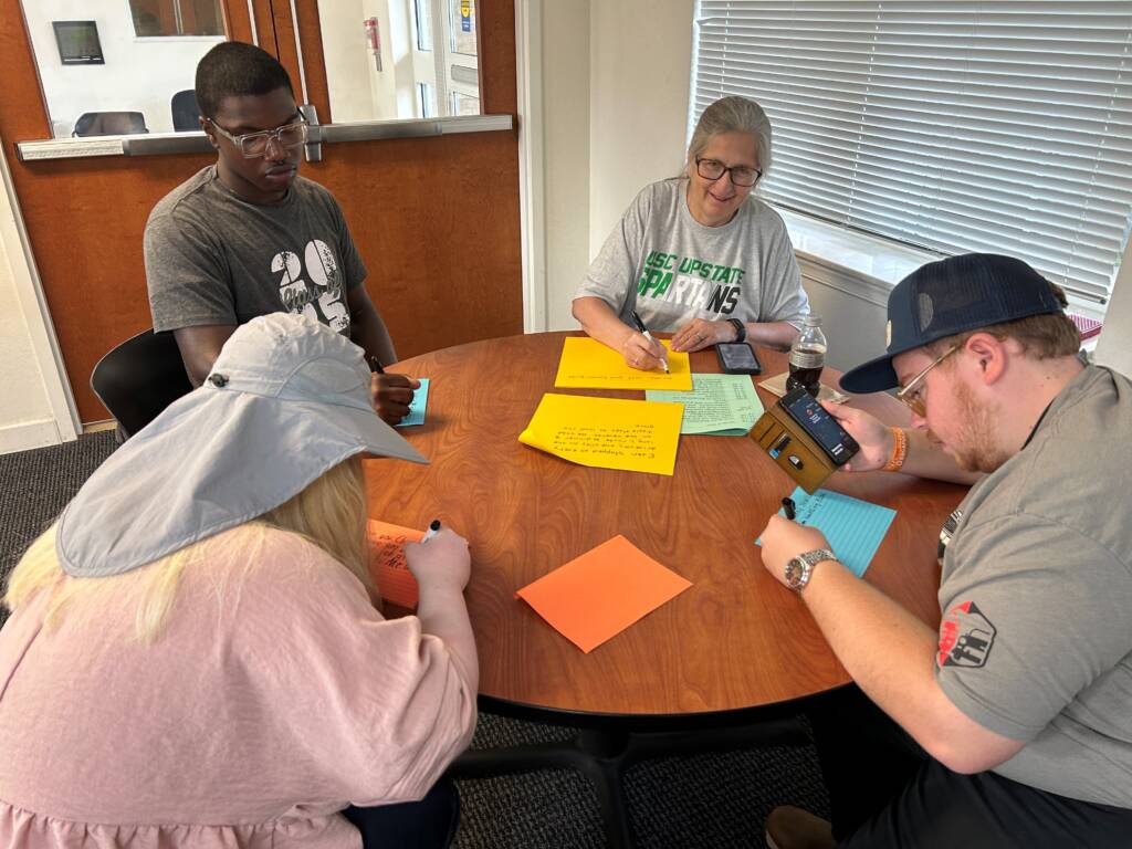 Dr. Herzberg and three travelers sit at a table writing posts for the wall. 