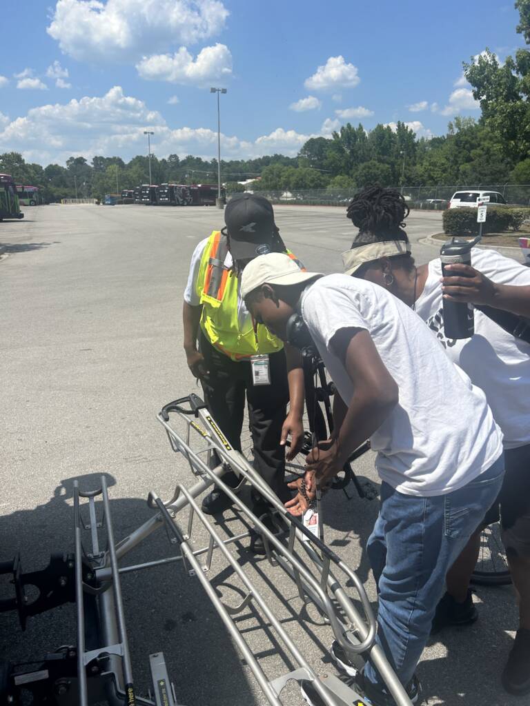 A male traveler explores a bike rack on the front of a fixed route bus as an employee gives directions. 