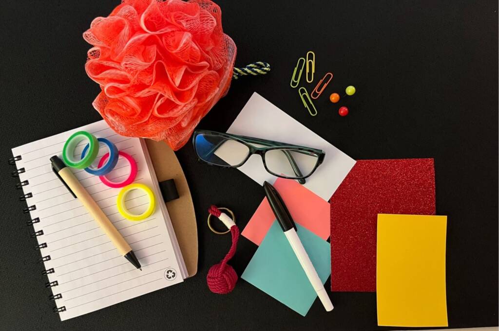 A desk top filled with various papers, a couple pens, glasses, and a bright orange scrunchy.